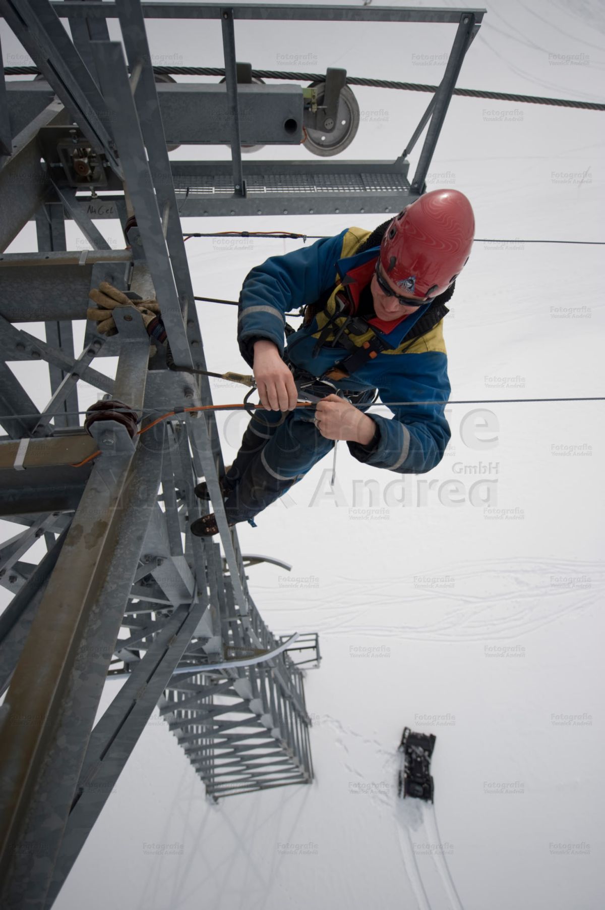 Bild zu dem Thema: Mitgliedschaften / Partner der Yannick Andrea GmbH. Das BIld zeigt ein Seilbahnmonteur bei der Arbeit auf einem Masten. Am Fuss des Sesselliftmastens parkiert das Schneemobil.